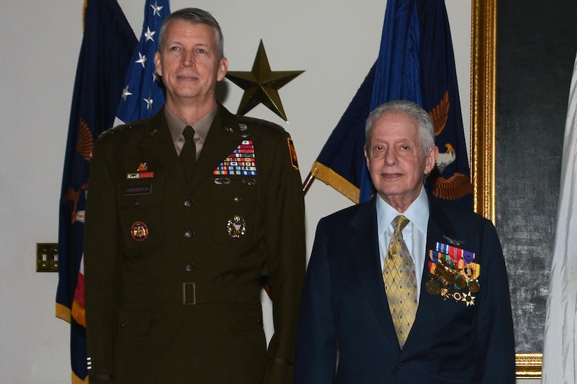 Two people stand next to one another with flags in the background. One is a uniformed service member. The other is wearing military insignia on their lapel.