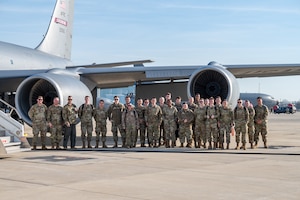 A man in a flight uniform reaches, pointing to a window on the underside of a plane. At right, a group of men and women in camo uniforms look at the window.