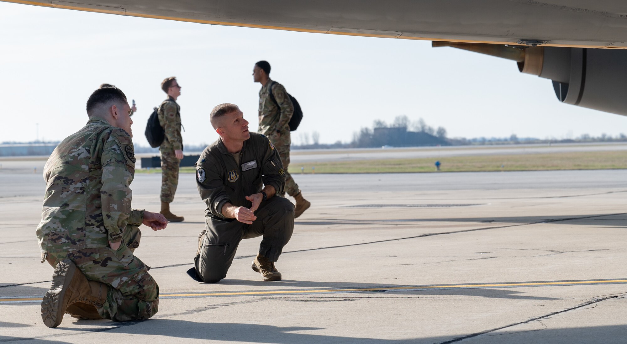 A man in a flight suit kneels underneath an aircraft while speaking to another man, in a military uniform, who is also kneeling. In the far background, two men in military uniform speak to one another.