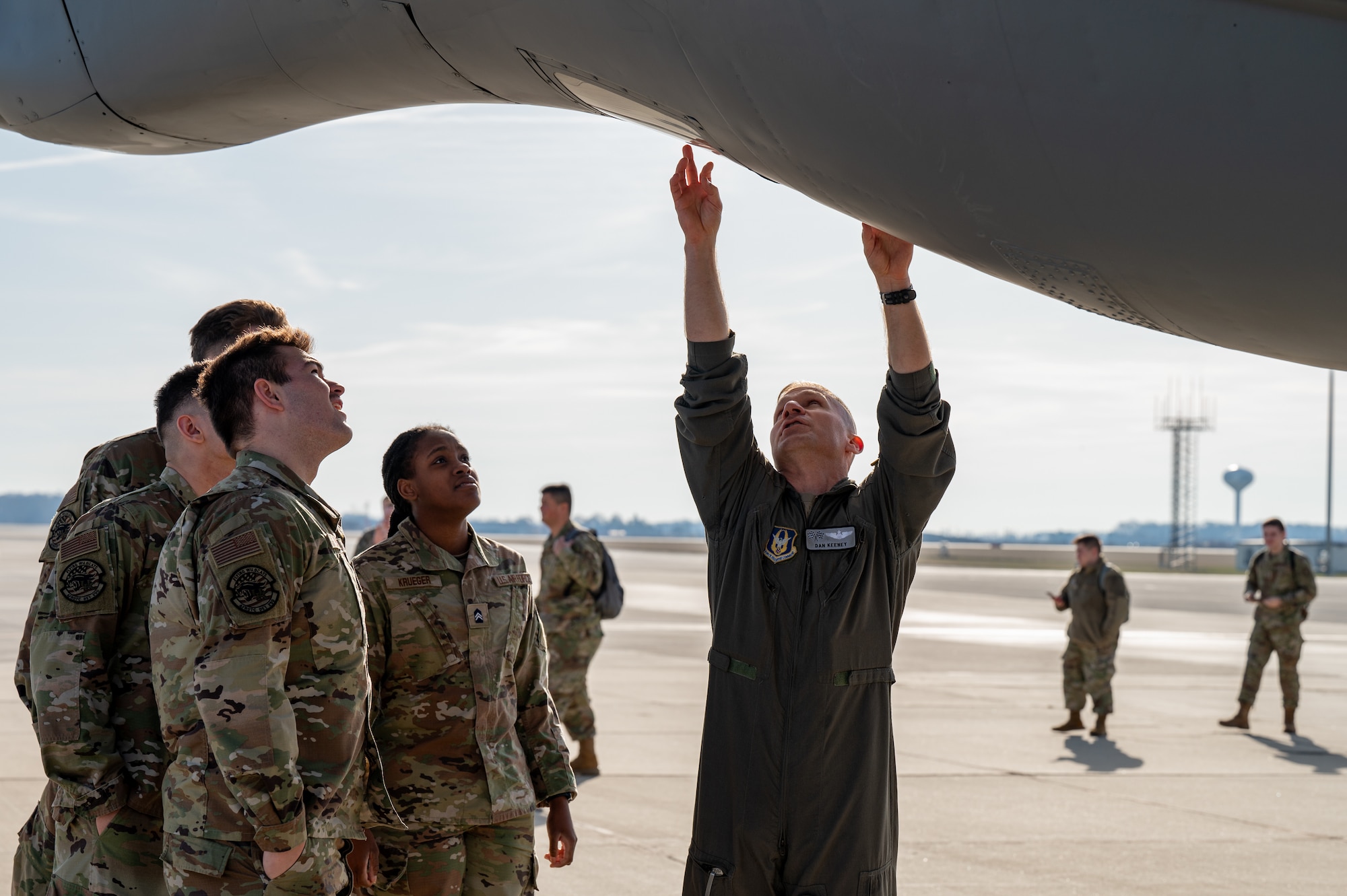 A man in a flight uniform reaches, pointing to a window on the underside of a plane. At right, a group of men and women in camo uniforms look at the window.