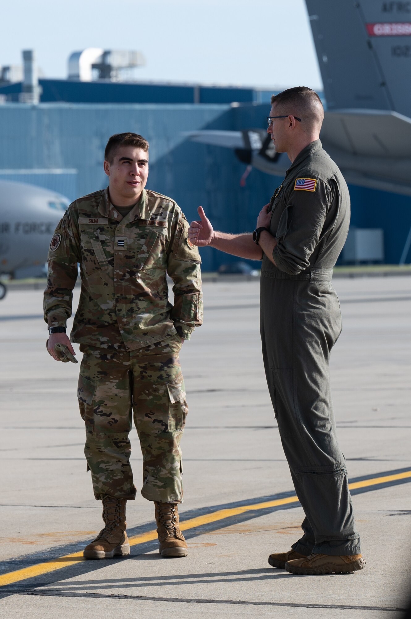 A man in a flight uniform gestures while talking to another man in a camouflage uniform. In the background, two military planes.
