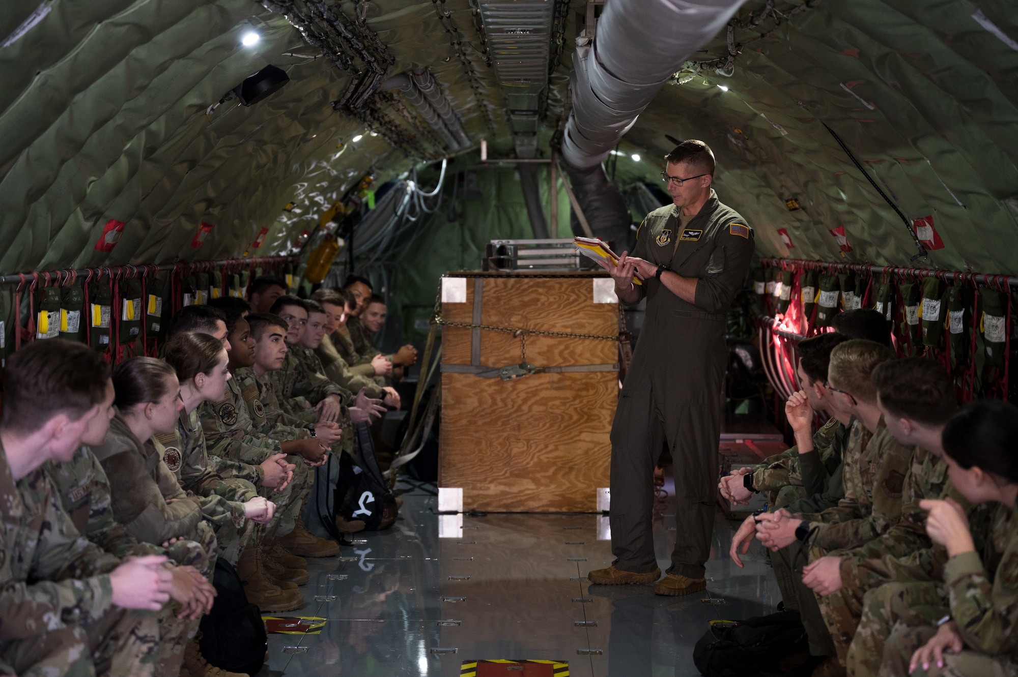 A man in a flight suit stands between two rows of men and women in military uniforms. He is speaking to them while holding a manual.