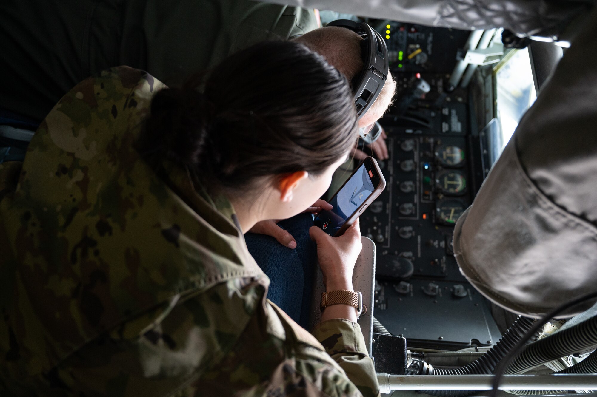 A woman in a military uniform holds a cell phone. On her screen, a white plane is visible.