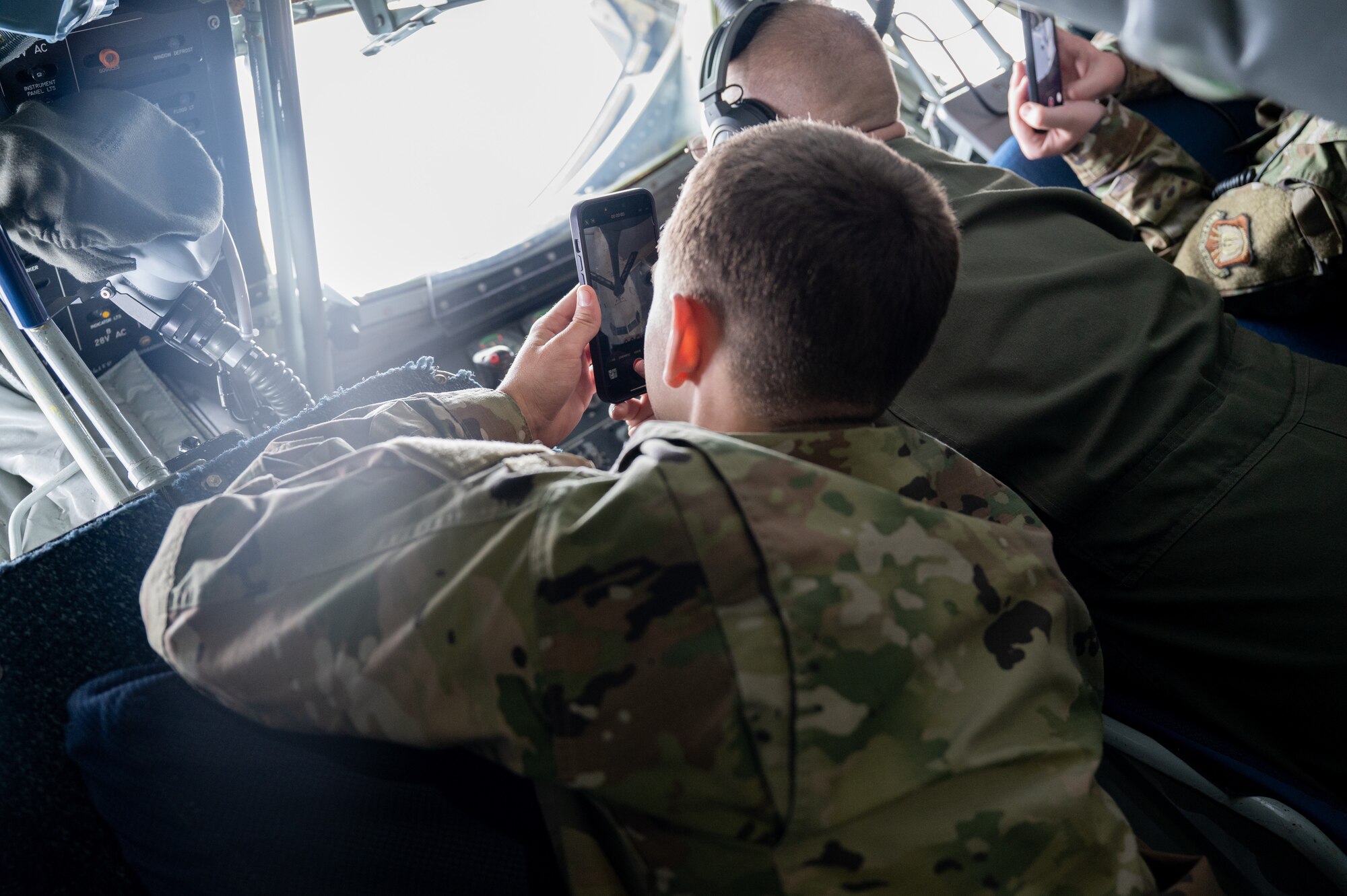 A man in a military uniform laying on his stomach holding a cell phone. On his screen, an airplane is visible.