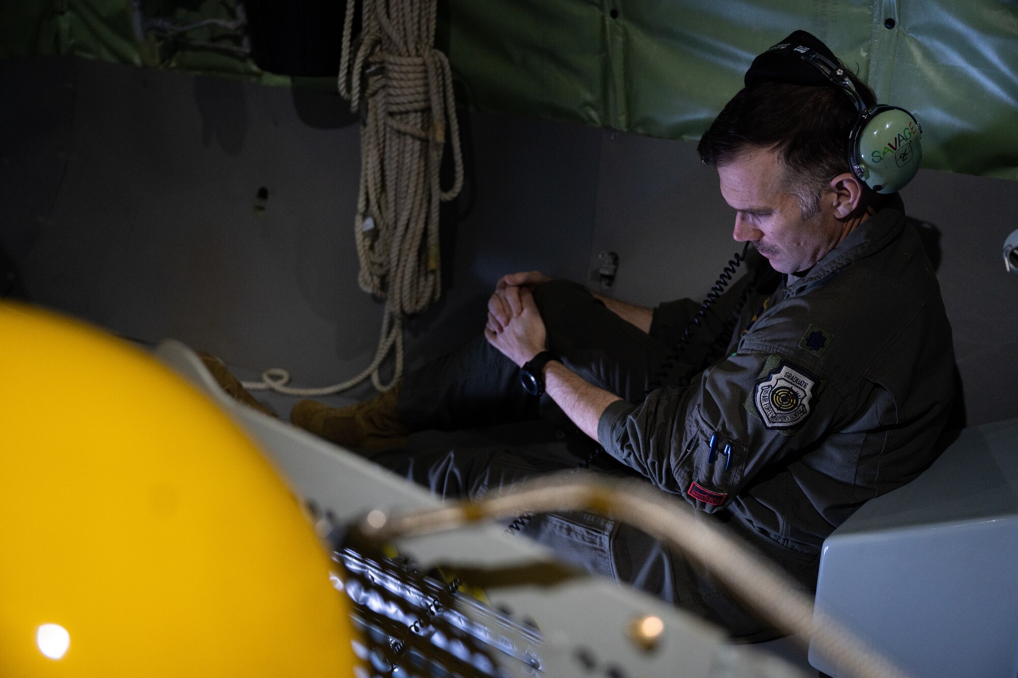 A man in a flight suit sits with one knee up, his hands on his propped knee. In the foreground, a yellow object that is blurry.