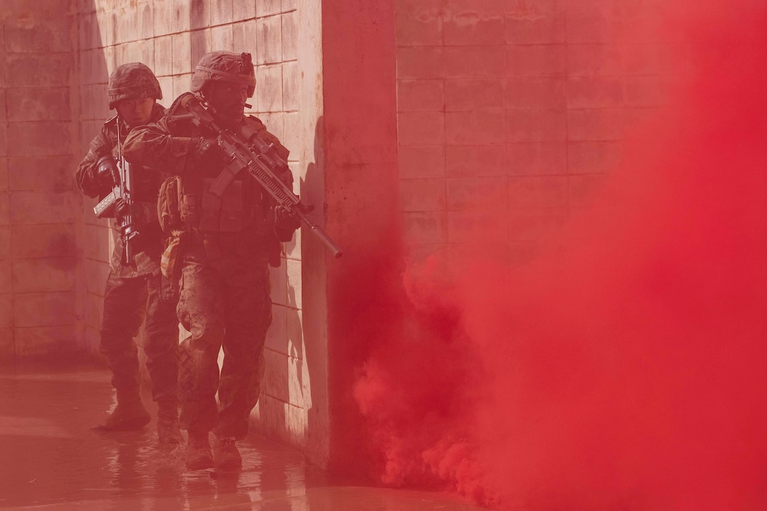 A U.S. and a South Korean marine in tactical gear standing next to a cement structure move towards clouds of red smoke.