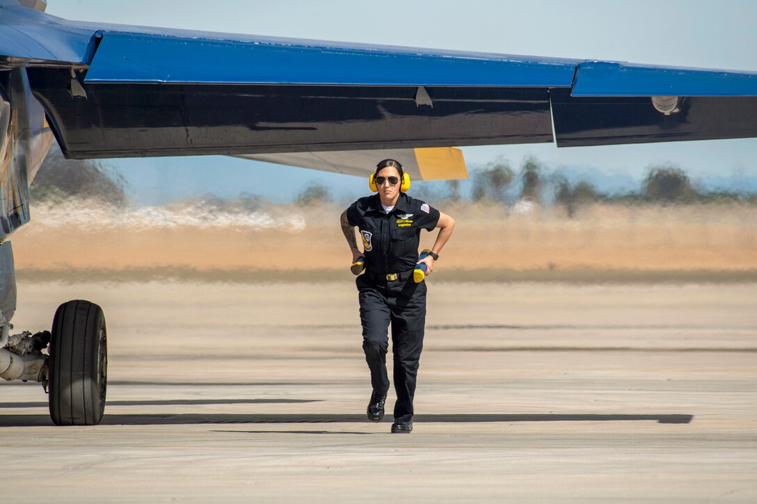 A service member carrying batons at their hips runs under the wings of an aircraft on a tarmac.