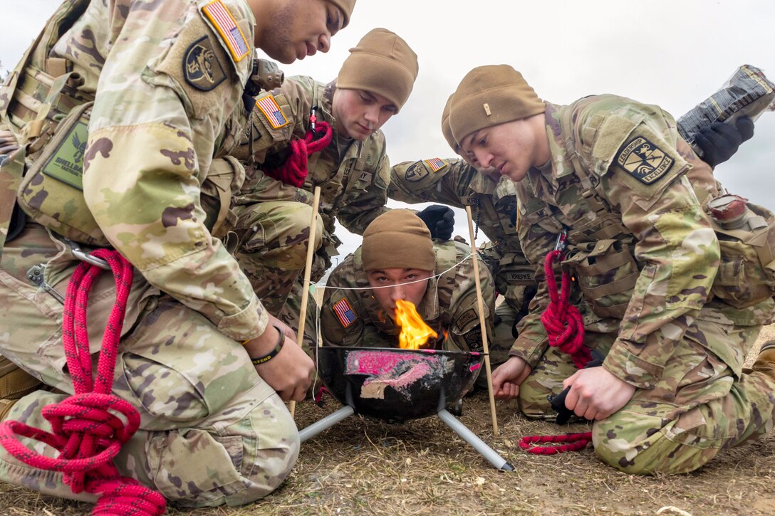 Army cadets with red bungee cords clipped to their uniforms gather around a small fire in a field.