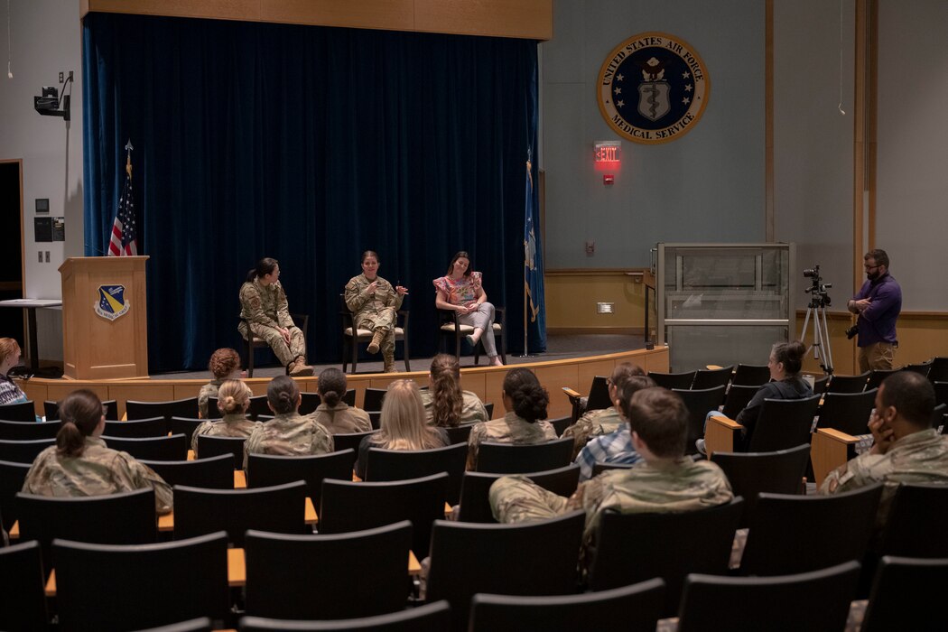 Three females sit on an auditorium stage while speaking to an audience of military and civilian personnel.