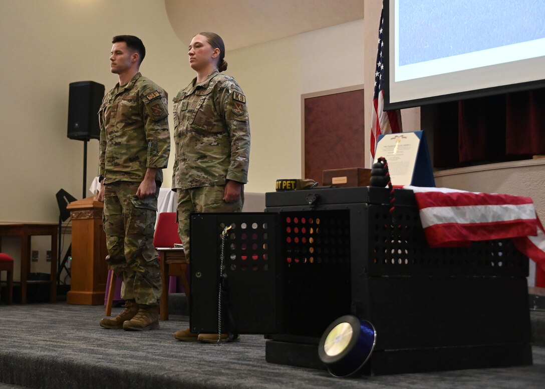 Two service members in uniform stand on a stage together in front of a military working dog's cage.