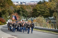 Army Soldiers in dark ceremonial uniforms carrying various musical instruments are marching up a paved street. There are white tombstones in rows on the right side of the picture and multicolored trees in the background.