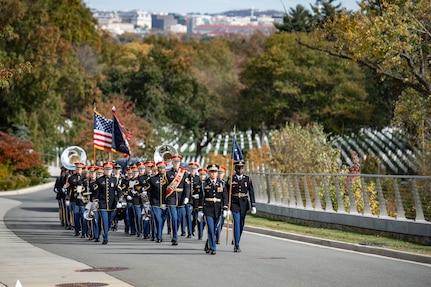 Army Soldiers in dark ceremonial uniforms carrying various musical instruments are marching up a paved street. There are white tombstones in rows on the right side of the picture and multicolored trees in the background.