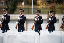 Army soldiers wearing dark ceremonial uniforms and carrying ceremonial rifles with bayonets affixed in fron of their bodies are matching into place in a cemetery that has all white marble tombstones.