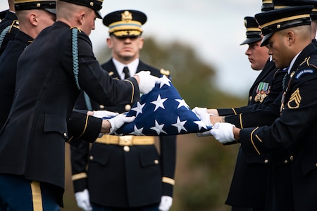 Army Soldiers wearing dark ceremonial uniforms and hats are folding the US flag as another soldier stands at attention nearby.