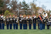 Army Soldiers in dark ceremonial uniforms are performing on musical instruments while standing on a green lawn. The director has a large, red sash across his torso.