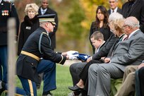 An Army Soldier wearing a dark ceremonial uniform and white gloves is handing a folded US flag to several family members of a fallen veteran. They are seated in green chairs and dressed in various colors of suits.
