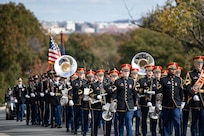 Army soldiers dress in dark ceremonial uniforms are marching in formation, in rows, while carrying various musical instruments. Behind them, soldiers are carrying ceremonial rifles with bayonets affixed over their right shoulders.