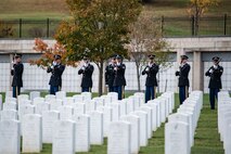 Soldiers in dark ceremonial uniforms are firing rifles in a cemetery with rows of white marble, rounded top headstones.