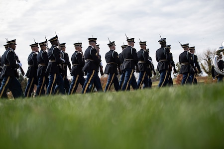 Army soldiers dress in dark ceremonial uniforms are marching in formation, in rows, away from the camera to the right, while carrying ceremonial rifles with bayonets affixed over their right shoulders. There is a large green lawn in the foreground.