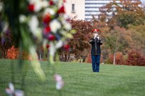A soldier in a dark ceremonial uniform is playing a trumpet while standing on a large grass lawn. In the forground are flowers.