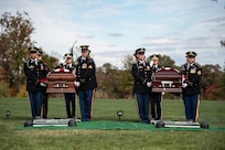 Army soldiers in dark ceremonial uniforms are carrying two flag-draped caskets across a green lawn towards their separate graves, which are covered with green cloth and metal frames for lowering the casket into the ground.