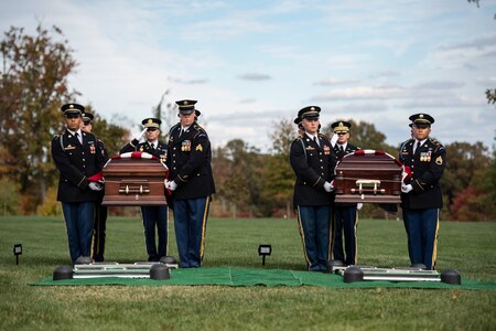 Army soldiers in dark ceremonial uniforms are carrying two flag-draped caskets across a green lawn towards their separate graves, which are covered with green cloth and metal frames for lowering the casket into the ground.