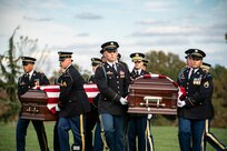 Army soldiers in dark ceremonial uniforms are carrying two flag-draped caskets across a green lawn.