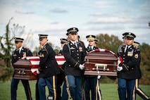 Army soldiers in dark ceremonial uniforms are carrying two flag-draped caskets across a green lawn.