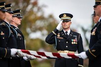 Army Soldiers wearing dark ceremonial uniforms and hats are folding the US flag as another soldier salutes.