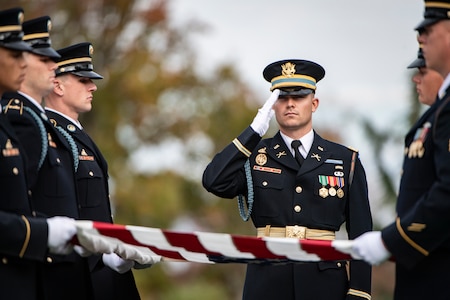 Army Soldiers wearing dark ceremonial uniforms and hats are folding the US flag as another soldier salutes.