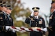 Army Soldiers wearing dark ceremonial uniforms and hats are folding the US flag as another soldier salutes.