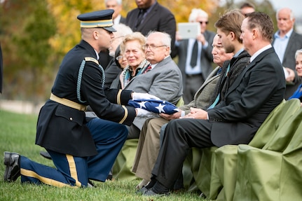 An Army Soldier wearing a dark ceremonial uniform and white gloves is handing a folded US flag to several family members of a fallen veteran. They are seated in green chairs and dressed in various colors of suits.