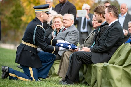 An Army Soldier wearing a dark ceremonial uniform and white gloves is handing a folded US flag to several family members of a fallen veteran. They are seated in green chairs and dressed in various colors of suits.