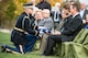 An Army Soldier wearing a dark ceremonial uniform and white gloves is handing a folded US flag to several family members of a fallen veteran. They are seated in green chairs and dressed in various colors of suits.