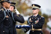 An Army soldier in a dark ceremonial uniform and hat is holding a folded US Flag out in front of him while another Soldier is saluting the flag. The each are wearing white gloves. Another soldier in the foreground is standing at attention.