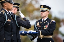 An Army soldier in a dark ceremonial uniform and hat is holding a folded US Flag out in front of him while another Soldier is saluting the flag. The each are wearing white gloves. Another soldier in the foreground is standing at attention.