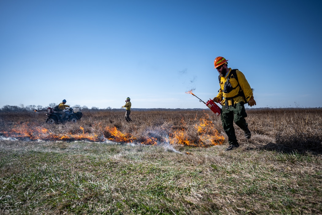 Adam Dietsch carries a fire drip torch with a trail of fire behind him from left to right. A firefighter and ATV with driver are in the background beyond the fire trail.