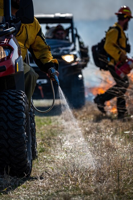A arm reaches out of an ATV with a hose spraying the grass, there is a firefighter in the background and another ATV.