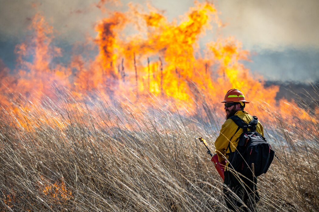 Adam Dietsch stands in a dry grassy field in the foreground with a massive flame in the background