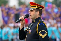 A soldier wearing a dark ceremonial uniform with bright red hat is holding a microphone and singing. In the background is a crown standing on bleachers.