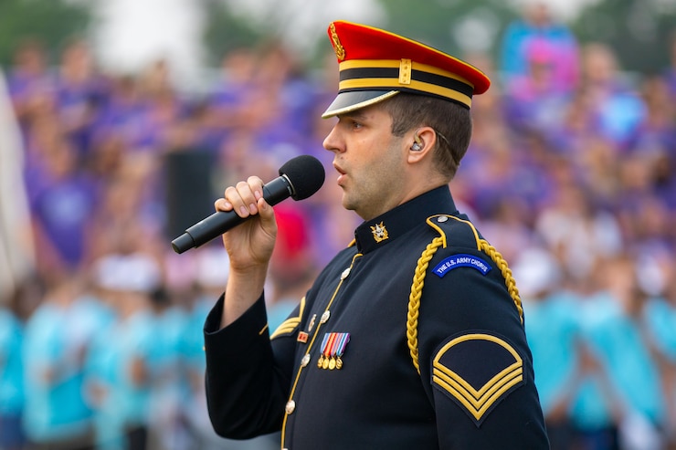 A soldier wearing a dark ceremonial uniform with bright red hat is holding a microphone and singing. In the background is a crown standing on bleachers.