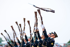 Soldiers dressed in dark ceremonial uniforms (dark coats with gold buttons, dark wheel hats and white gloves) are tossing rifles into the air. The rifles have silver bayonets affixed.