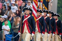 Soldiers dressed in Revolutionary War-era uniforms (dark blue with red trim coats, cream-colored pants and dark with white trim tri-colored hats) are marching toward the camera carrying a mix of rifles and flags. In the background are audience members.