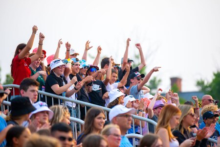 People in various colors of clothes are standing on silver colored bleachers with their arms up.