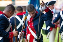 Soldiers dressed in Revolutionary War era uniforms (blue with red trim coats and cream-colored pants and white criss-cross straps across their torsos) are adjusting their uniforms.
