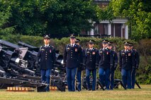 About 8 Army soldiers in dark ceremonial uniforms are marching into place near large black cannons on a green lawn.