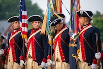 Soldiers wearing Revolutionary War-era uniforms (dark blue with red trim coats, dark blue with white trim tri-cornered hats, and cream-colored vests and pants) are carrying flags and rigles, and they are looking in various directions as they make adjustments to their formation.