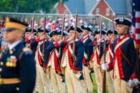 Soldiers wearing Revolutionary War-period uniforms (dark blue with red trim coats, cream-colored pants and dark blue with white trim tri-cornered hats) are adjusting the rifles that they are carrying against their shoulders. In the foreground is a soldier in a modern Army dark ceremonial uniform (dark jacket with gold buttons and dark wheel hat)