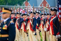 Soldiers wearing Revolutionary War-period uniforms (dark blue with red trim coats, cream-colored pants and dark blue with white trim tri-cornered hats) are adjusting the rifles that they are carrying against their shoulders. In the foreground is a soldier in a modern Army dark ceremonial uniform (dark jacket with gold buttons and dark wheel hat)