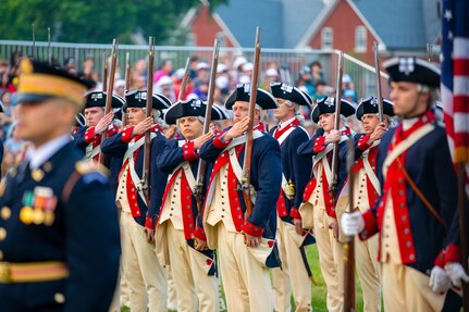 Soldiers wearing Revolutionary War-period uniforms (dark blue with red trim coats, cream-colored pants and dark blue with white trim tri-cornered hats) are adjusting the rifles that they are carrying against their shoulders. In the foreground is a soldier in a modern Army dark ceremonial uniform (dark jacket with gold buttons and dark wheel hat)